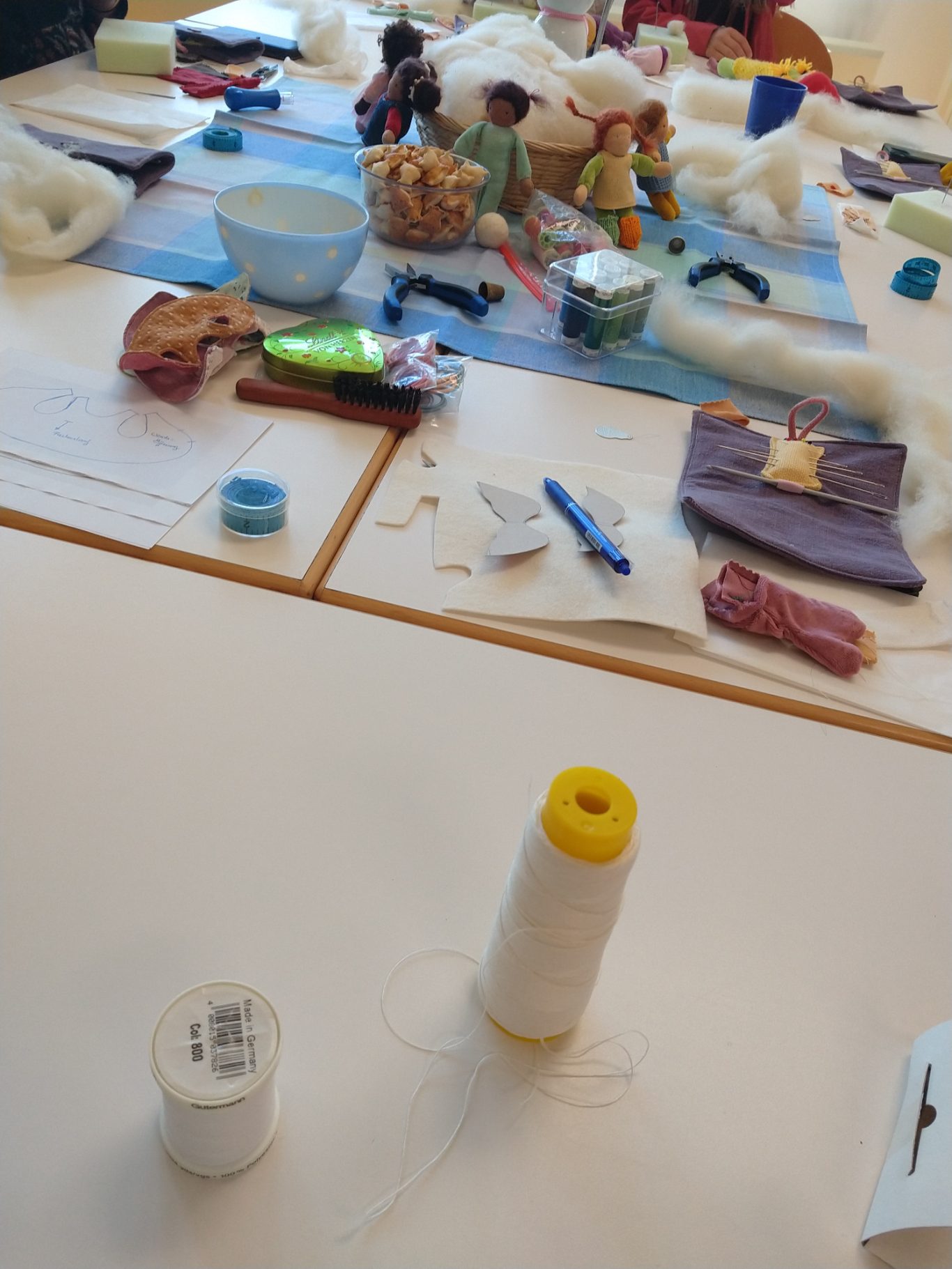 Man sieht einen Tisch mit verschiedenen Nähutensilien, die zum Puppenmachen benötigt werden. Außerdem sitzen im Hintergrund ein paar Puppen