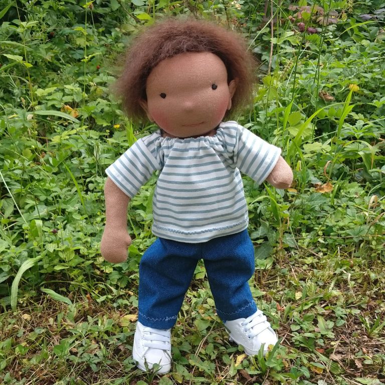 Quinn von Susis Puppenglück Stoffpuppenjunge mit Dysmelie am linken Arm. Er hat dunkle Haare und trägt ein geringeltes T-Shirt, Jeans und Sneaker.