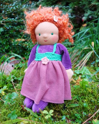Man sieht eine Schmusepuppe mit lila Nicky-Körper und roten Locken. Sie trägt ein Kleid mit grünem Oberteil, Blüte und Tüllrock