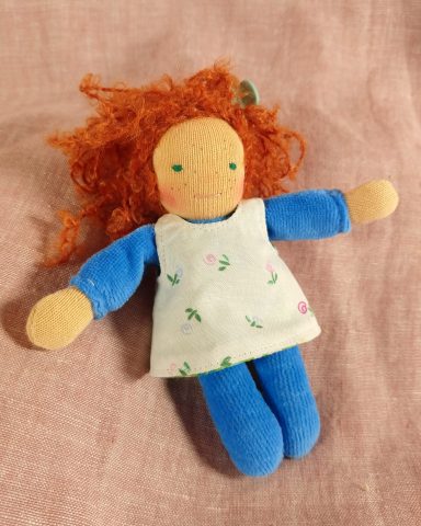 Man sieht eine Taschenpuppe mit blauem Nicky-Körper und roten Locken. Sie trägt ein weißes Kleid mit kleinen Blümchen.