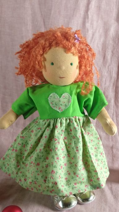 Das Foto zeigt ein stehendes Stoffpuppenmädchen mit rotlockigem Haar. Sie trägt ein grünes Kleid mit Herz-Applikation und Streublümchen.