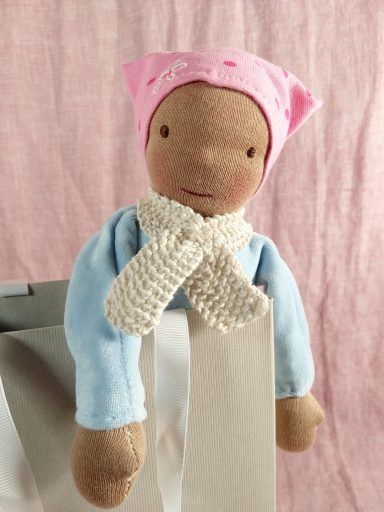Man sieht eine Schmusepuppe mit helbaluem Nicky-Körper. Sie trägt eine gepunktete rosa Mütze und hat einen weißen Schal um den Hals.