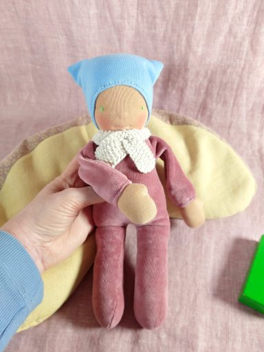 Man sieht eine Schmusepuppe mit altrosa Nicky-Körper. Sie trägt eine hellblaue Mütze und hat einen weißen Schal um den Hals.