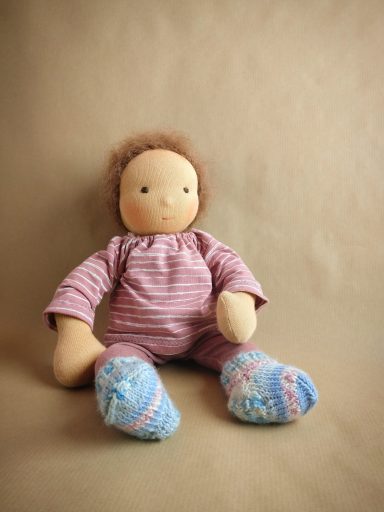 Puppenbaby mit braunen Wuschelhaaren und braunen Augen. Es trägt eine gestrickte Socken, rosa Leggins und ein geringeltes Oberteil.