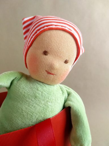Schmusepuppe mit hellgrünem Nicky-Körper und rot-weiß geringelter Mütze. Sie steht in einer Tasche und ist daher nur bis zur Brust zu sehen.