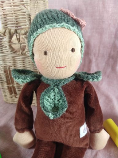 Man sieht eine Schmusepuppe mit braunem Nicky-Körper. Sie trägt eine grüne Mütze mit rosa Blume und hat um den Hals einige Blätter