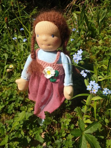 Man sieht eine Schmusepuppe mit hellblauem Nicky-Körper und braunen Zöpfen. Sie trägt ein altrosa Kleid mit weißer Blüte