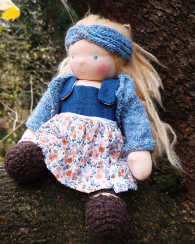 Man sieht eine Puppe mit blonden Haaren. Sie trägt ein Kleid mit Jeans-Oberteil und geblümten Rock. Darüber trägt sie eine Strickjacke