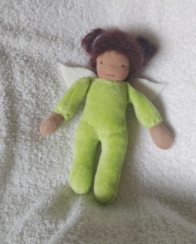 Man sieht eine Taschenpuppe mit grünem Nicky-Körper und braunen Haarschnecken. Sie hat am Rücken kleine Flügel