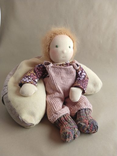 Puppenbaby mit roten Wuschelhaaren und grünen Augen. Es trägt eine gestrickte, rosa Latzhose und ein geblümtes Oberteil