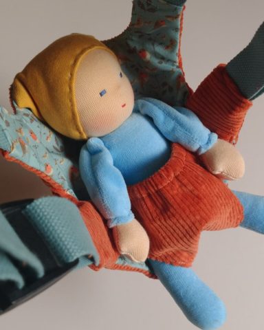 Man sieht eine Schmusepuppe mit hellblauem Nicky-Körper und gelber Mütze, die in einer Puppentrage sitzt
